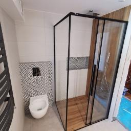 Usługi Budowlano Remontowe Michał Powęska - Nowoczesna łazienka z kabiną prysznicową z czarnymi ramami i drewnopodobną podłogą, wisząca toaleta z geometrycznym wzorem na ścianie, grzejnik drabinkowy po lewej stronie.