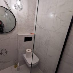 Usługi Budowlano Remontowe Michał Powęska - Nowoczesna łazienka z marmurowymi płytkami, wiszącą toaletą z rolką papieru, okrągłym lustrem i geometryczną lampą wiszącą; widoczne akcesoria do czyszczenia podłogi.