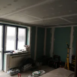 Wnętrze w trakcie montażu płyt kartonowo-gipsowych, widoczne płyty na ścianach i suficie, zaszpachlowane łączenia, okno zabezpieczone folią malarską, narzędzia i worki z zaprawą na podłodze.