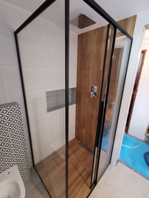 Nowoczesna kabina prysznicowa typu walk-in z czarnymi profilami, deszczownicą i drewnopodobną okładziną ściany, z widocznym odpływem liniowym w podłodze.