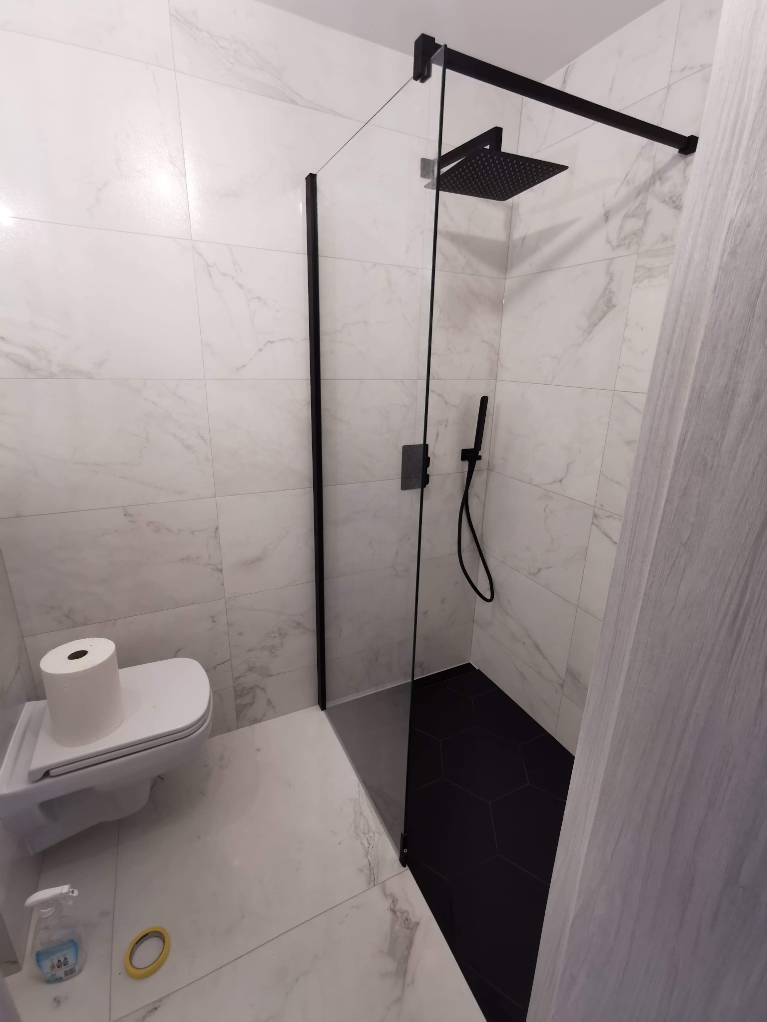 Nowoczesna łazienka z czarną armaturą, kabiną prysznicową typu walk-in z czarnymi profilami i deszczownicą, białe płytki imitujące marmur na ścianach i podłodze, czarne heksagonalne płytki...