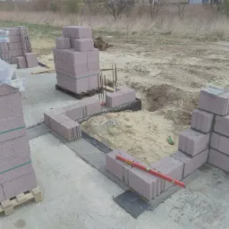 Wznoszenie ścian z bloczków betonowych na fundamencie; widoczne palety z bloczkami, poziomica i warstwa izolacji pionowej fundamentu.
