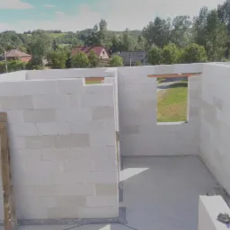 Wnętrze surowego budynku z białych bloczków, widoczne otwory okienne i wylana betonowa posadzka, na zewnątrz zieleń i zabudowania.