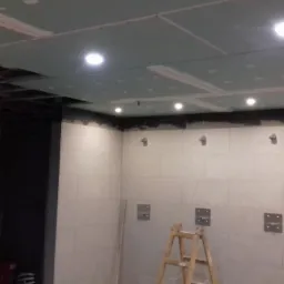 Montaż płyt karton-gips na suficie z wbudowanym oświetleniem punktowym. Widoczne płytki na ścianach i drabina. Prace wykończeniowe w pomieszczeniu.