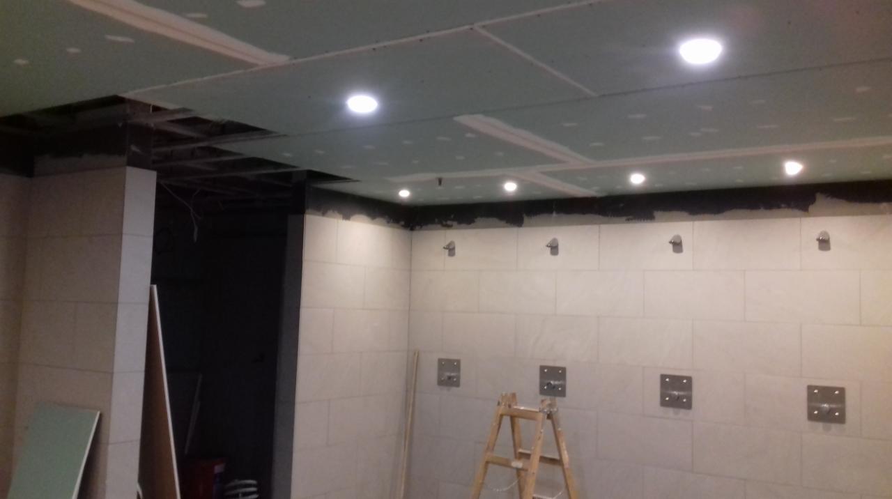 Montaż płyt karton-gips na suficie z wbudowanym oświetleniem punktowym. Widoczne płytki na ścianach i drabina. Prace wykończeniowe w pomieszczeniu.