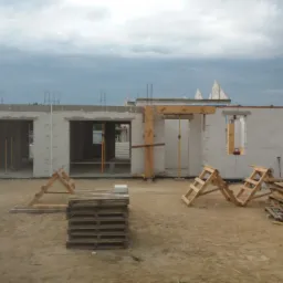 Surowy stan domu w trakcie budowy z białych bloczków, widoczne drewniane szalunki otworów okiennych i drzwiowych, palety i drabiny na piaszczystym podłożu, w tle pochmurne niebo.