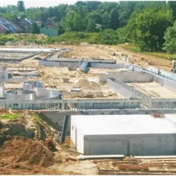 Budowa kompleksu zbiorników retencyjnych z betonu i stali, widok z góry na plac budowy z mini koparką w tle, herb miasta w lewym górnym rogu.