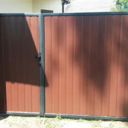 Brązowa furtka z paneli pionowych z czarną ramą i słupkami, zamontowana przy jasnym budynku z widoczną rynną, w tle zieleń i budynek gospodarczy.