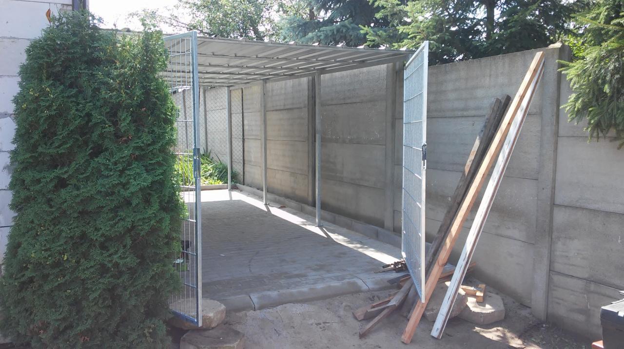 Metalowa konstrukcja z zadaszeniem i otwartą furtką z siatki, przylegająca do betonowego ogrodzenia, z tujami po lewej stronie i drewnianymi deskami opartymi o ogrodzenie.
