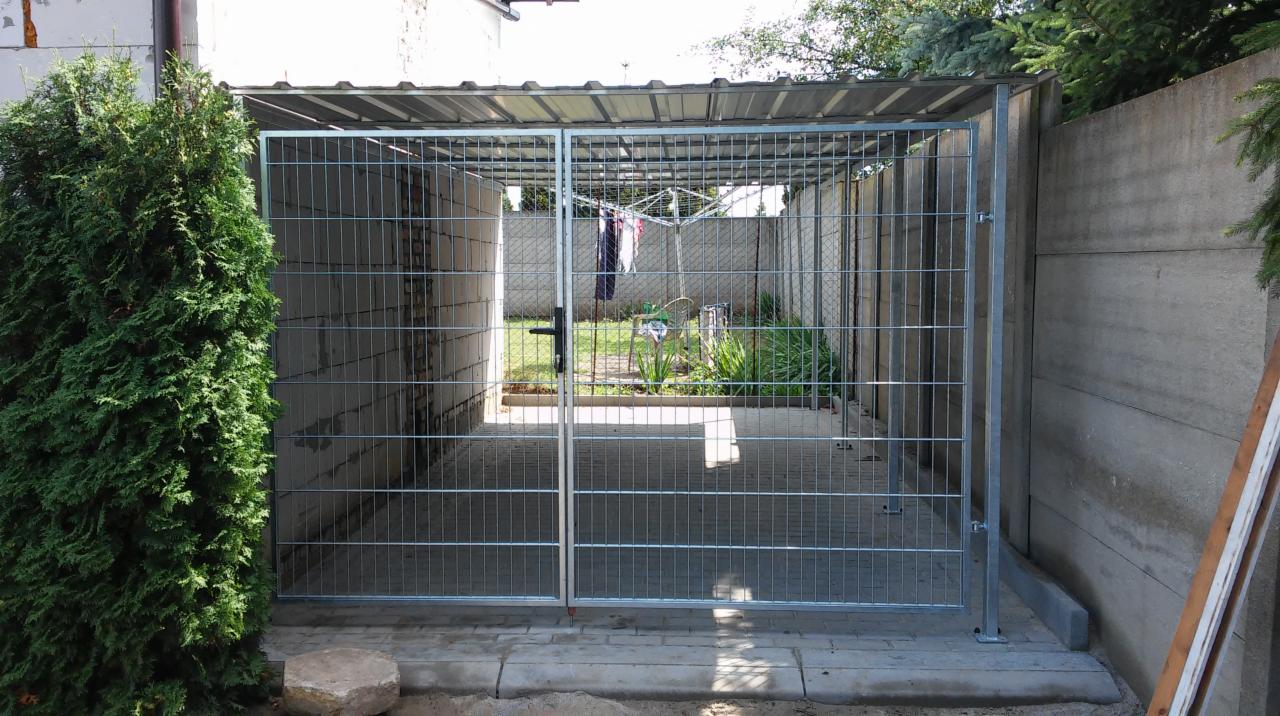 Metalowa konstrukcja z kratownicy i zadaszeniem, przypominająca kojec lub wiatę, z widoczną suszarką na pranie w tle, otoczona zielenią i betonowymi ścianami.