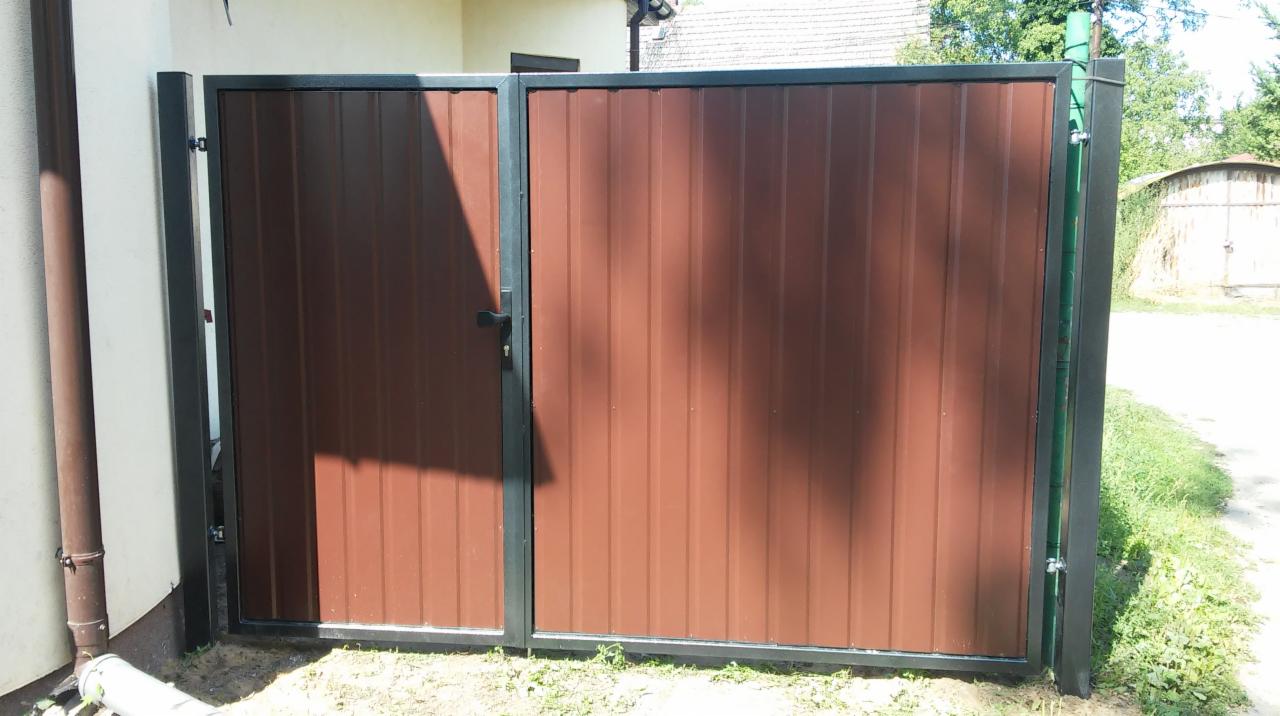 Brązowa furtka z paneli pionowych z czarną ramą i słupkami, zamontowana przy jasnym budynku z widoczną rynną, w tle zieleń i budynek gospodarczy.