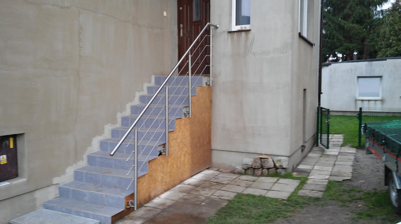 Schody zewnętrzne z szarych płytek z metalową balustradą prowadzące do drzwi wejściowych, z widocznym fragmentem elewacji budynku w trakcie wykończenia i tymczasowym zabezpieczeniem schodów z płyty...