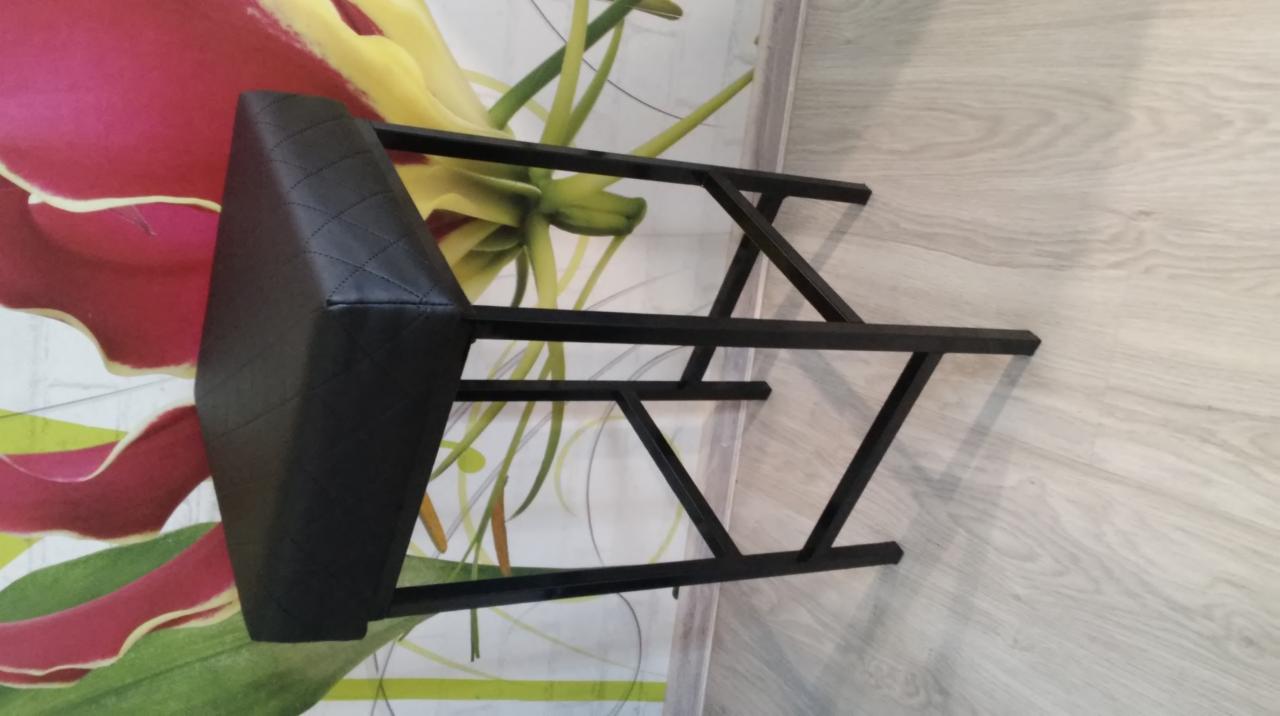 Nietypowe ujęcie nowoczesnego krzesła barowego z czarnym, pikowanym siedziskiem i metalową konstrukcją na tle wzorzystej ściany.