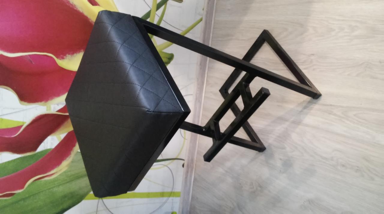 Nietypowy stołek barowy z czarnym, pikowanym siedziskiem i geometryczną, metalową podstawą, ustawiony na tle wzorzystej tapety z motywem kwiatowym i paneli podłogowych.