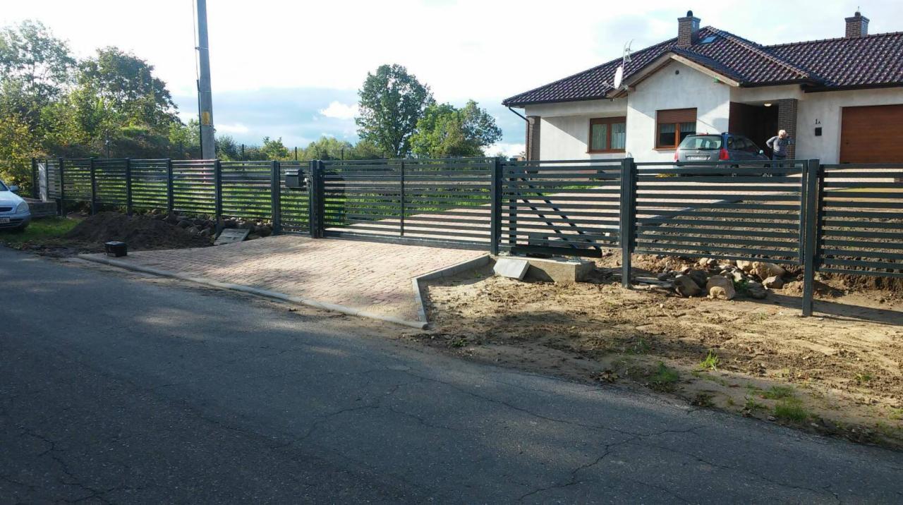 Nowoczesne, poziome ogrodzenie panelowe w kolorze grafitowym z bramą wjazdową na posesję z kostki brukowej, w tle dom jednorodzinny z brązową dachówką i samochodem