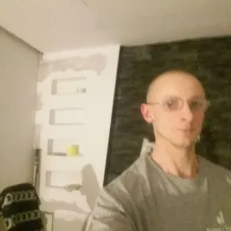 Mężczyzna w okularach pozuje na tle ściany z częściowo wykończonymi gładziami i elementami z płyty karton gips, widoczne niedoskonałości wykończenia.