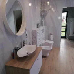 Nowoczesna łazienka z marmurowymi płytkami, owalnym lustrem nad drewnianą szafką z białą umywalką, wiszącą toaletą i bidetem, oraz drewnopodobną podłogą z widokiem na ogród przez czarne drzwi...