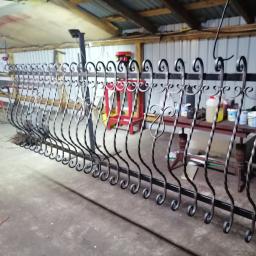 Patryk Andrzejewski - Częściowo ukończone, czarne, dekoracyjne ogrodzenie z kutego żelaza w warsztacie, widoczne narzędzia i farby na stole w tle.