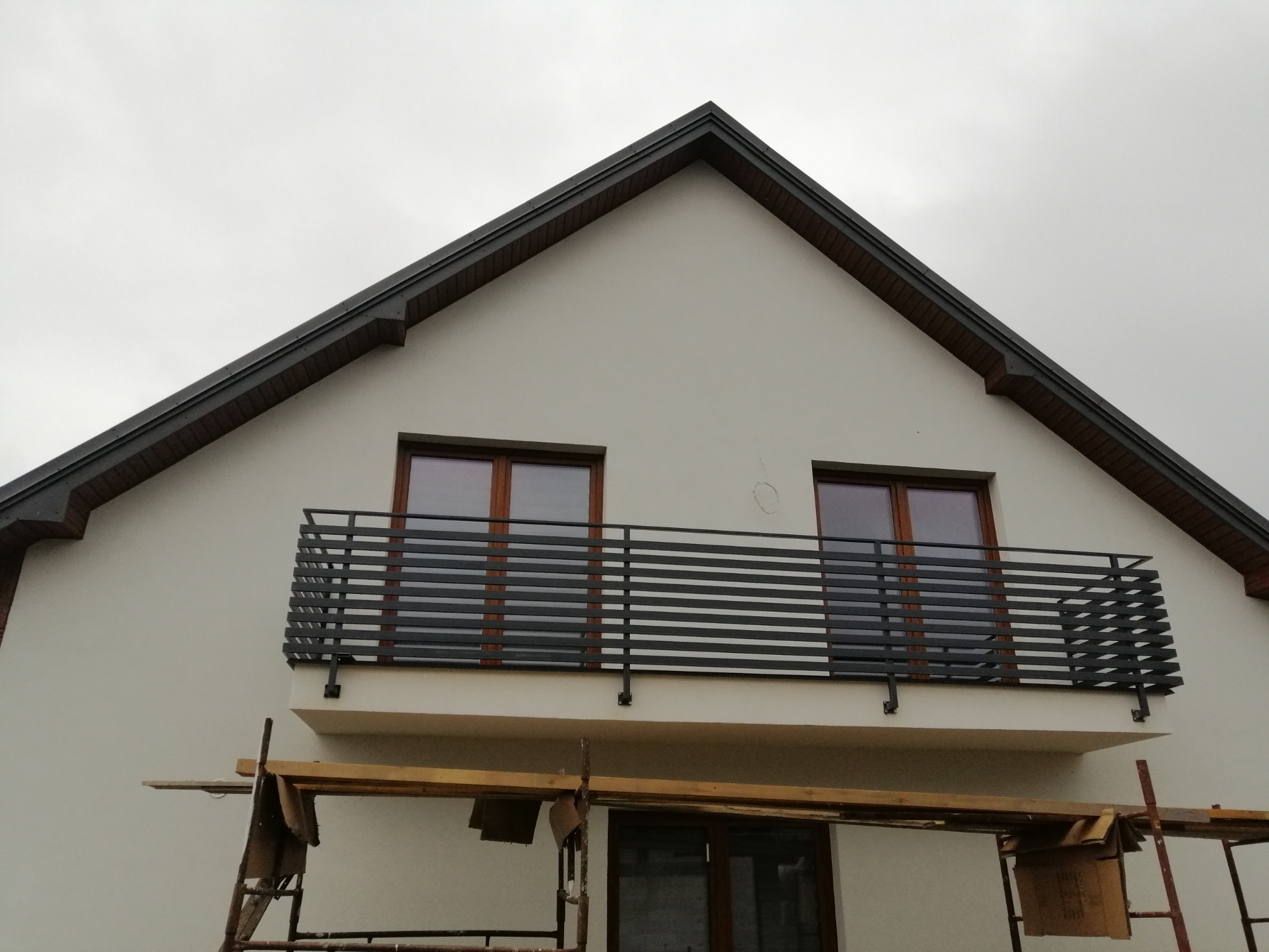 Nowoczesna balustrada balkonowa z metalowych poziomych profili, zamontowana w nowym budynku, widoczne rusztowanie sugerujące trwające prace wykończeniowe.