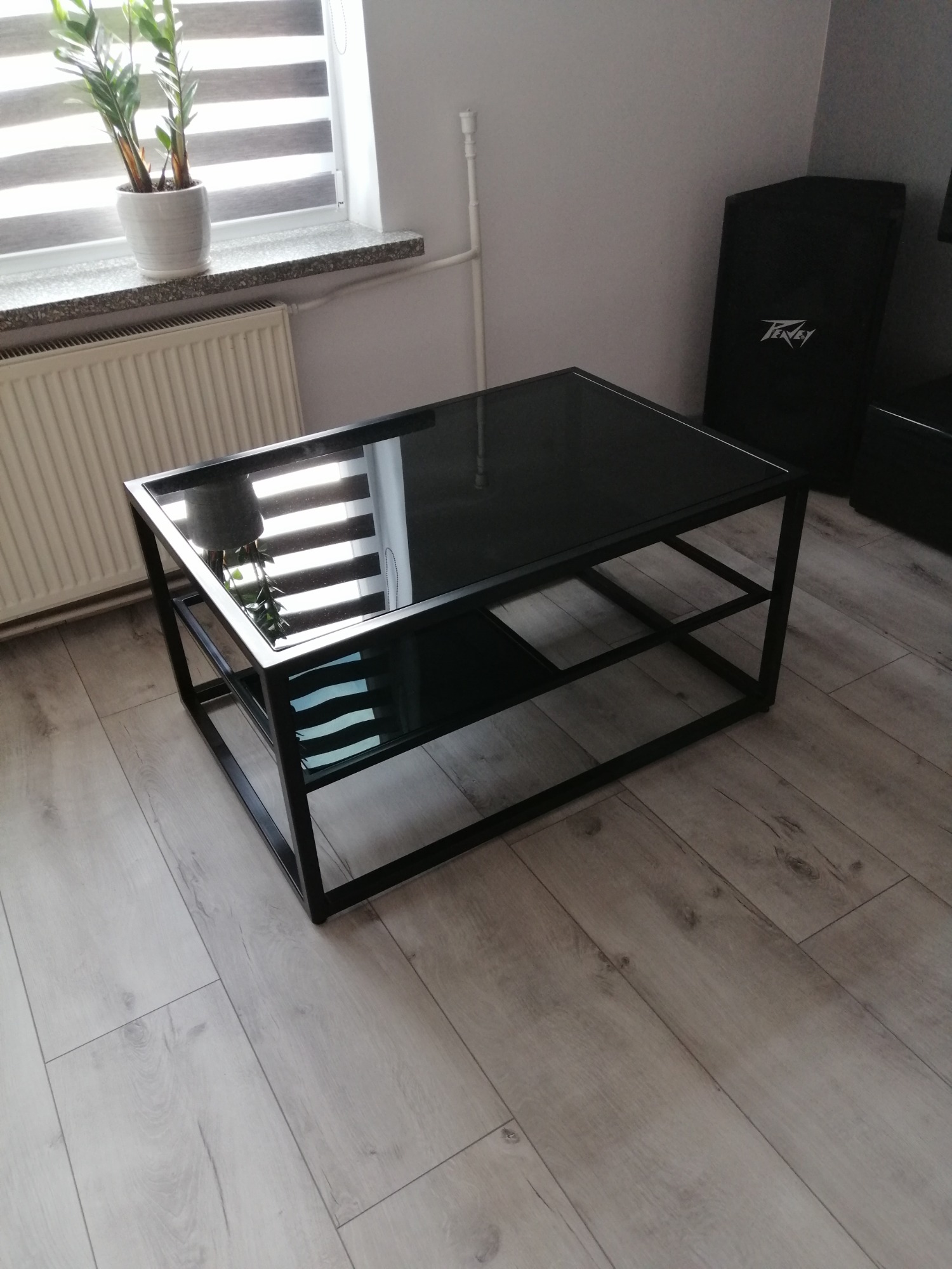 Czarny stolik kawowy z metalową ramą i szklanym blatem, stojący na panelach podłogowych, w tle widoczny grzejnik i roślina doniczkowa na parapecie.