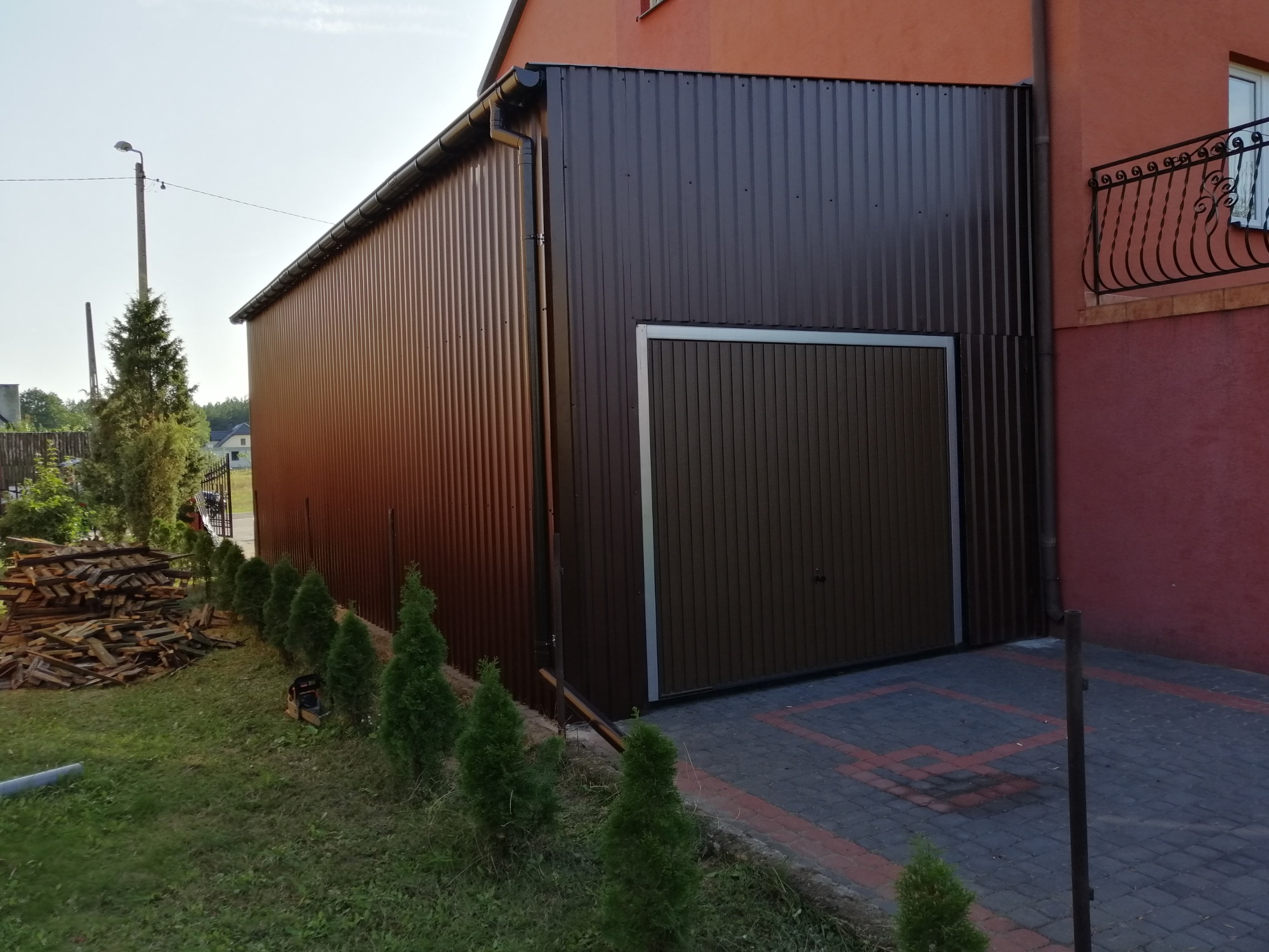Nowoczesny, brązowy garaż blaszany z bramą segmentową, przylegający do domu, widok z zewnątrz, z małym drzewkiem iglastym wzdłuż ściany