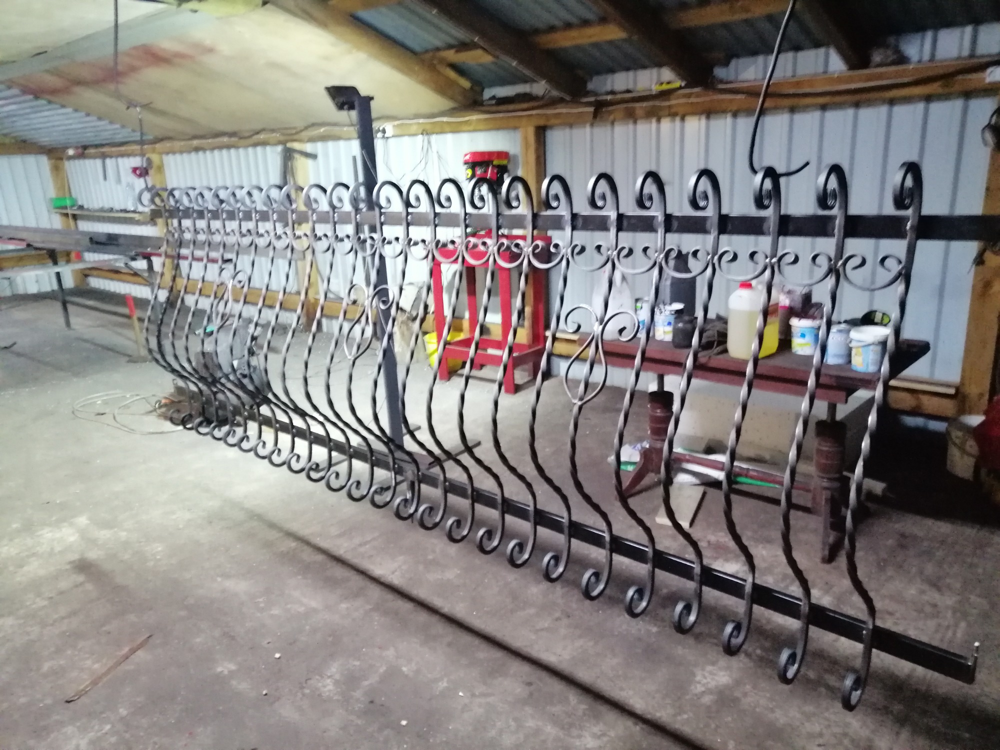 Częściowo ukończone, czarne, dekoracyjne ogrodzenie z kutego żelaza w warsztacie, widoczne narzędzia i farby na stole w tle.