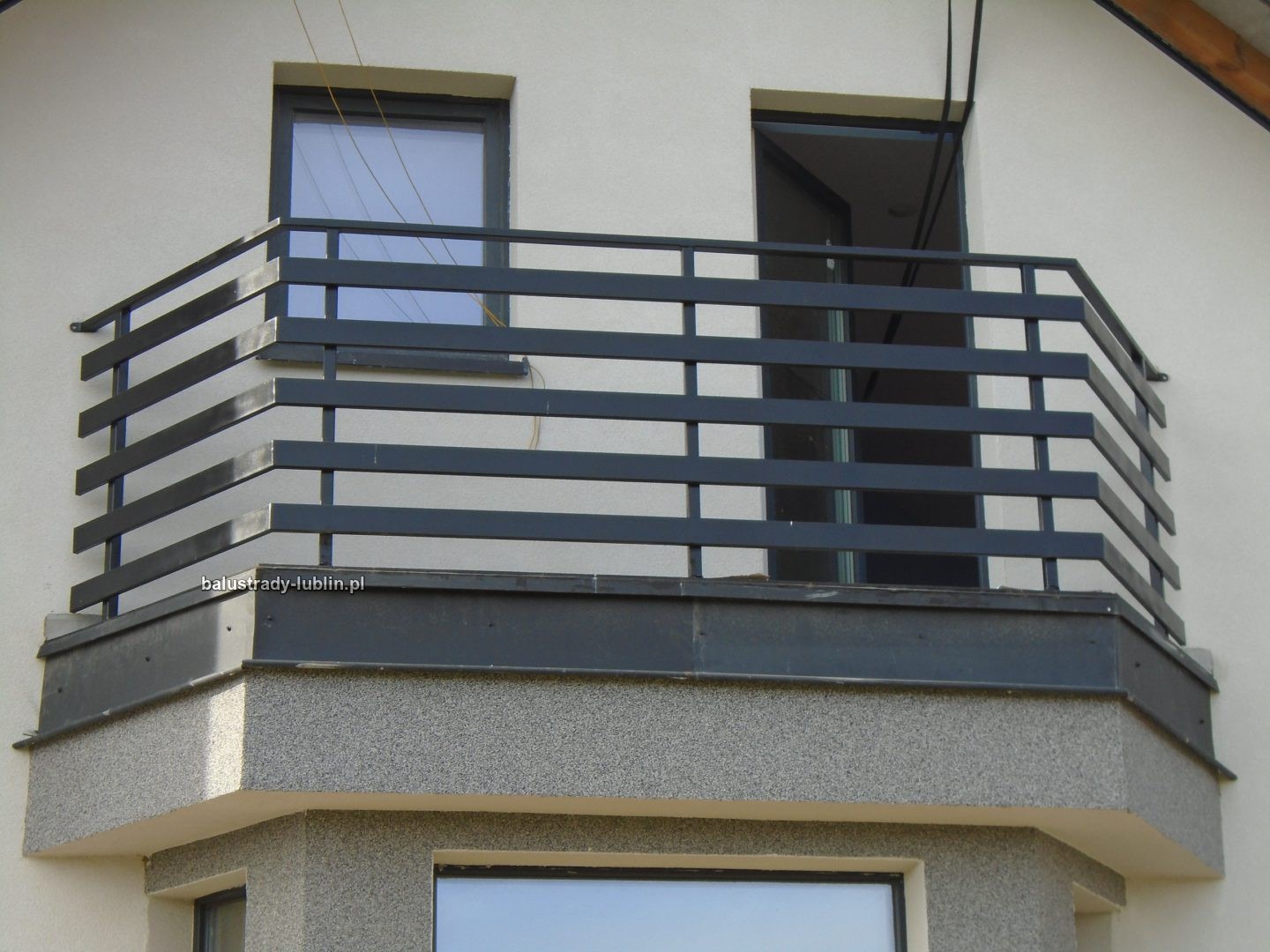 Nowoczesna, metalowa balustrada balkonowa z poziomymi profilami, pomalowana na grafitowo, zamontowana na balkonie z szarą, ziarnistą elewacją i widocznym oknem.
