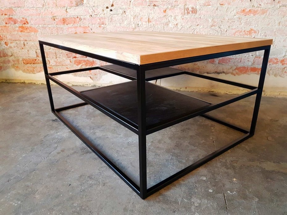 Stolik kawowy o kwadratowym blacie z jasnego drewna, osadzony na czarnej, metalowej konstrukcji z dwoma półkami, na tle ceglanej ściany.