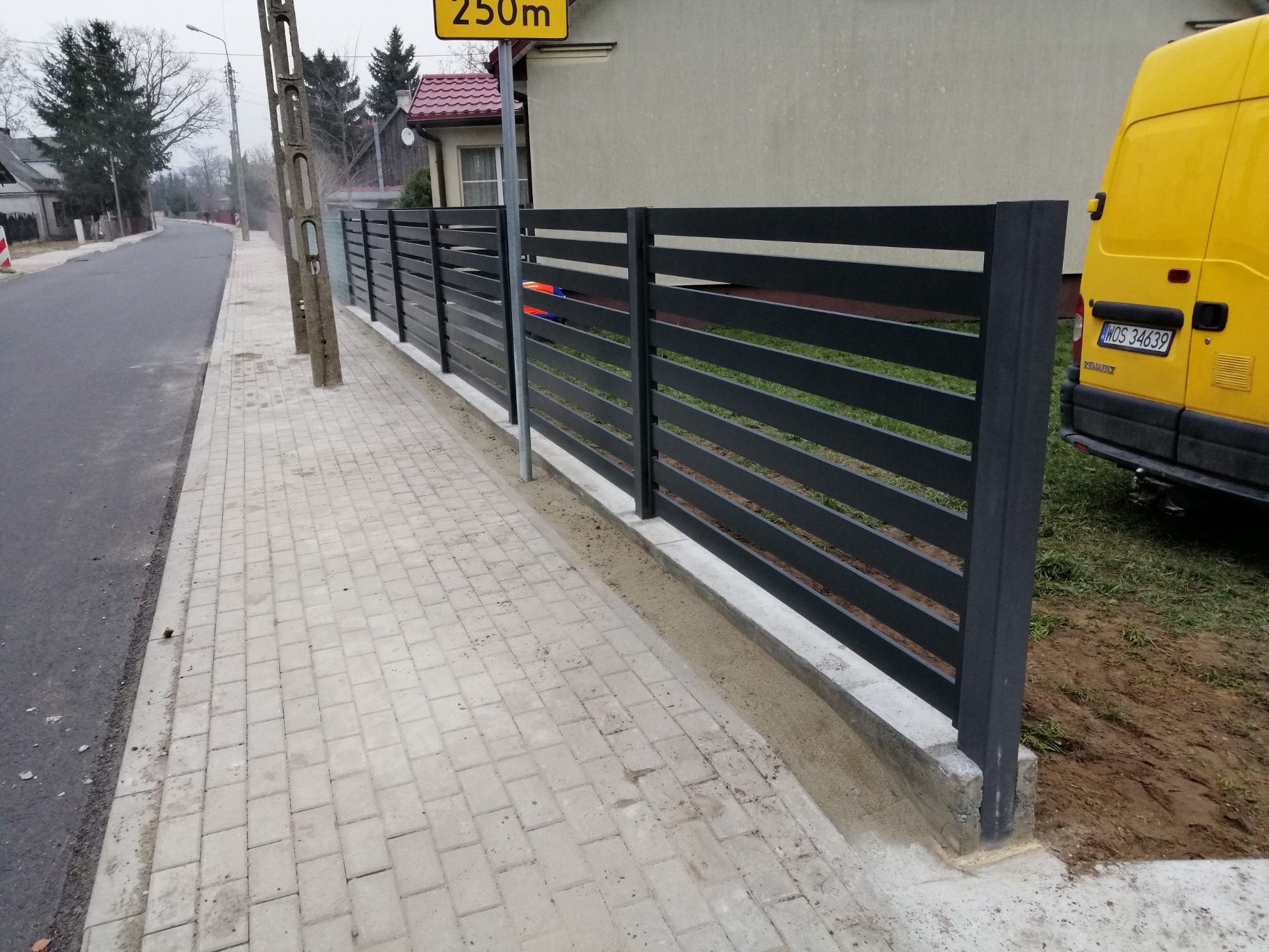 Nowoczesne, poziome przęsła w kolorze grafitowym, tworzące ogrodzenie posesji, widoczne z poziomu chodnika wyłożonego kostką brukową, z żółtym samochodem dostawczym w tle.