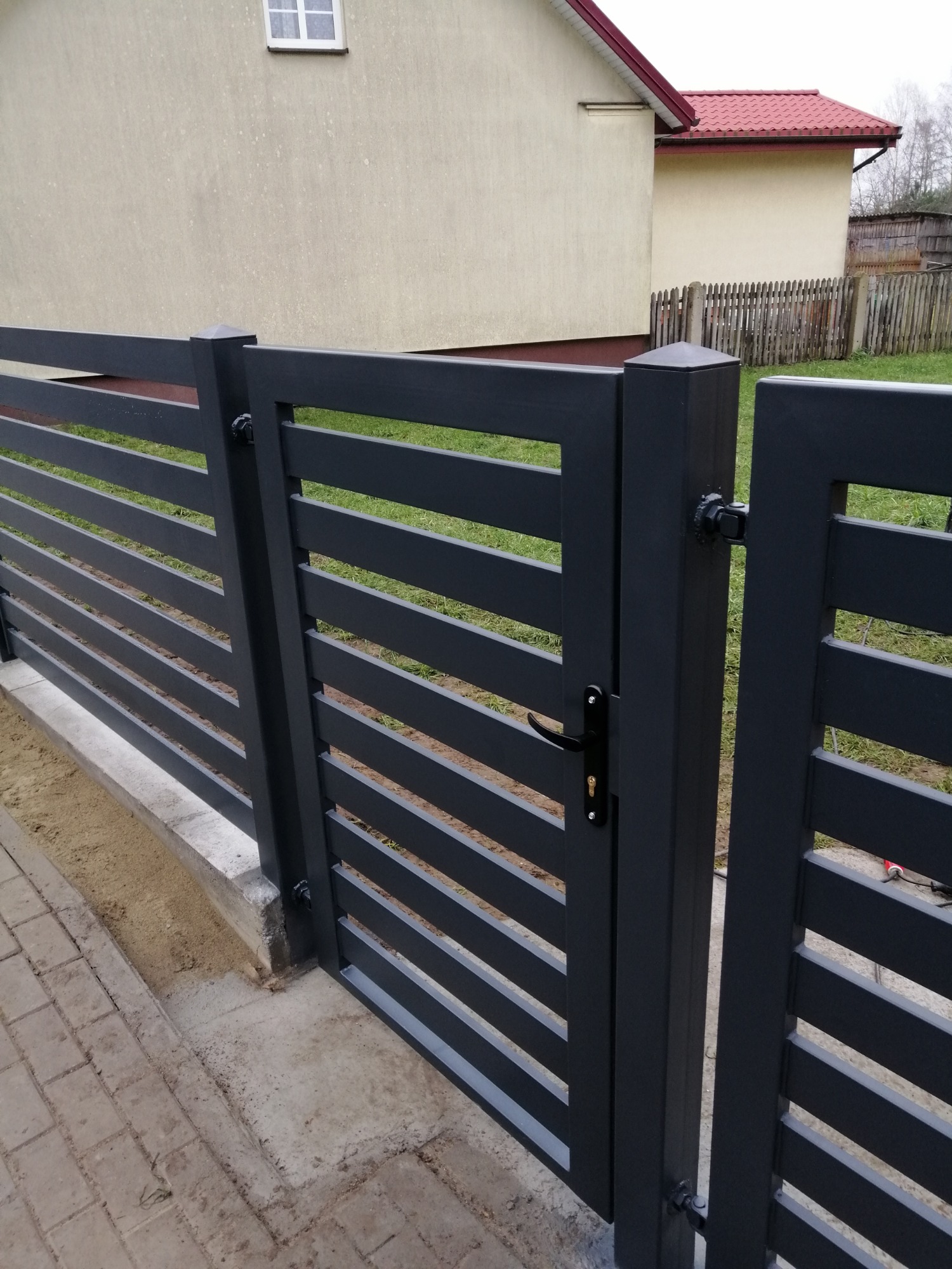 Nowoczesna, pozioma brama panelowa w kolorze antracytowym z czarną klamką, widoczna na tle trawnika i budynku z czerwoną dachówką.