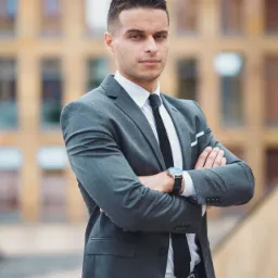 Młody mężczyzna w szarym garniturze, białej koszuli i czarnym krawacie, z zegarkiem na ręce, stoi na zewnątrz z założonymi rękami na tle budynku z wieloma oknami.