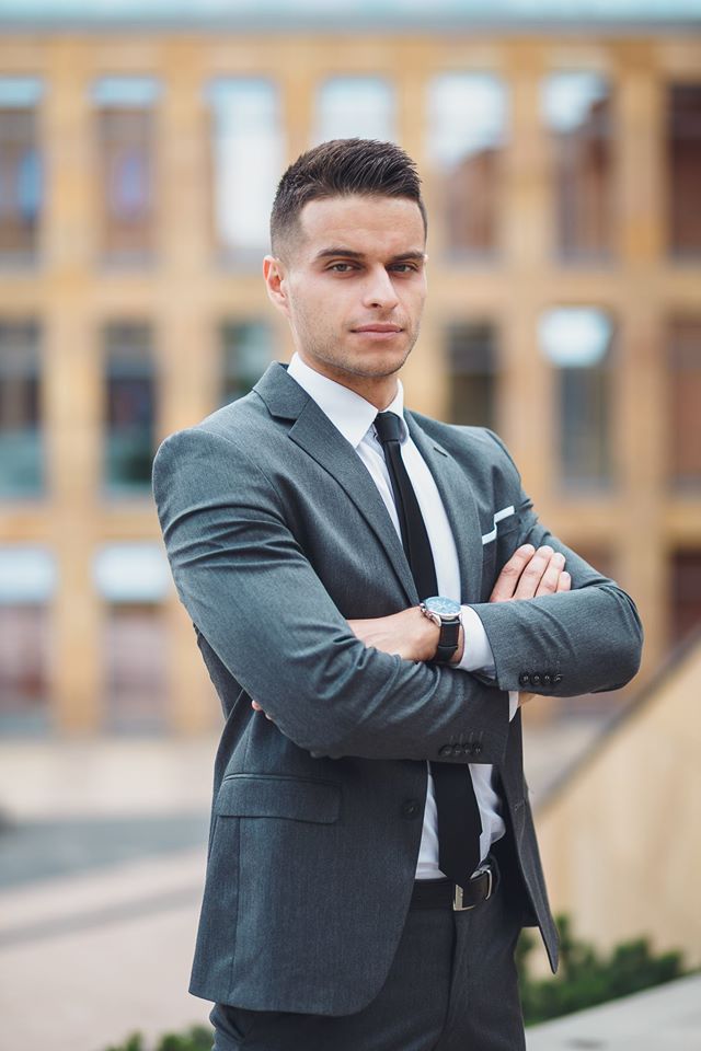 Młody mężczyzna w szarym garniturze, białej koszuli i czarnym krawacie, z zegarkiem na ręce, stoi na zewnątrz z założonymi rękami na tle budynku z wieloma oknami.