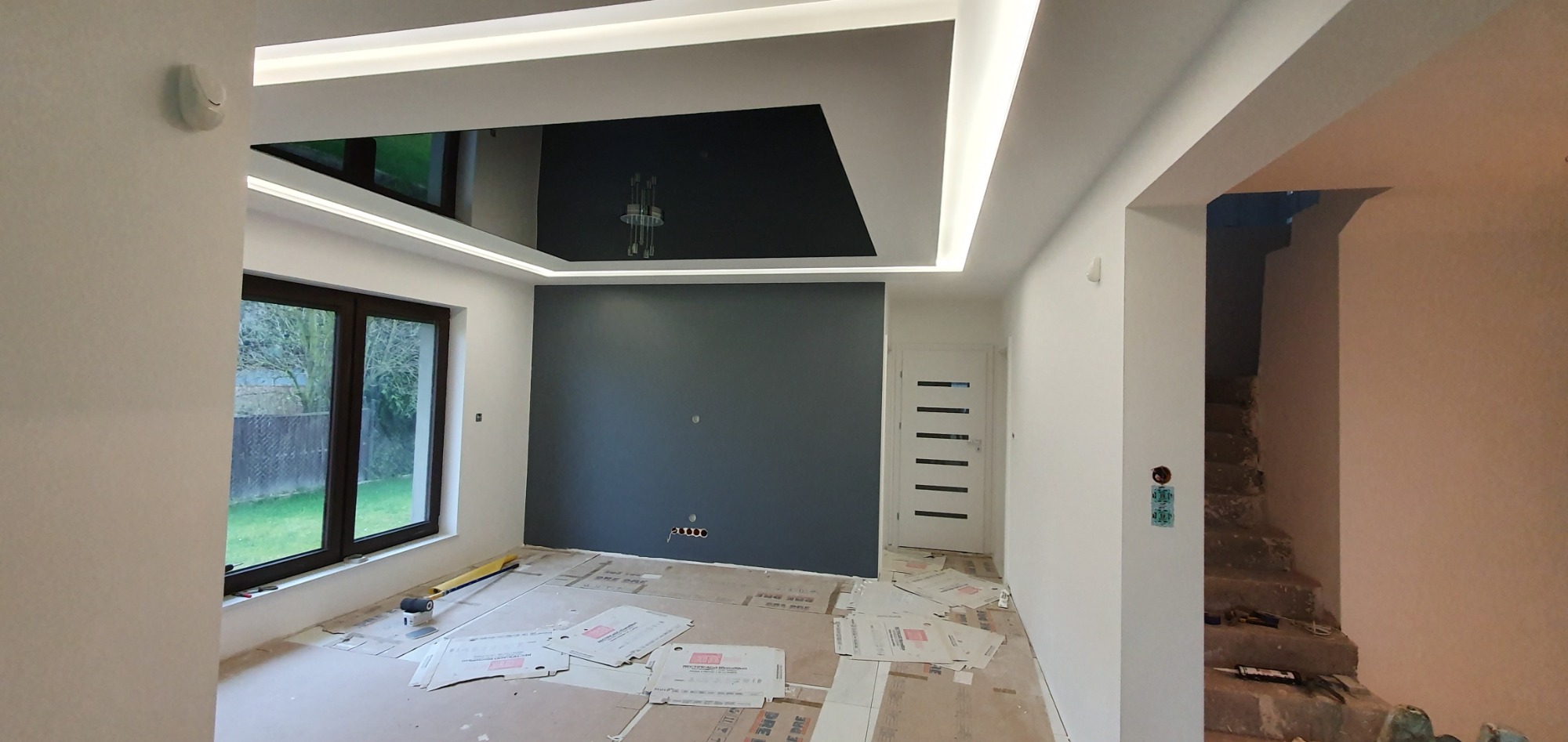 Pokój w trakcie odświeżania: pomalowana na szaro ściana, białe ściany boczne, czarny sufit podwieszany z oświetleniem LED, zabezpieczona podłoga, widoczne gniazdka elektryczne, okno z widokiem...