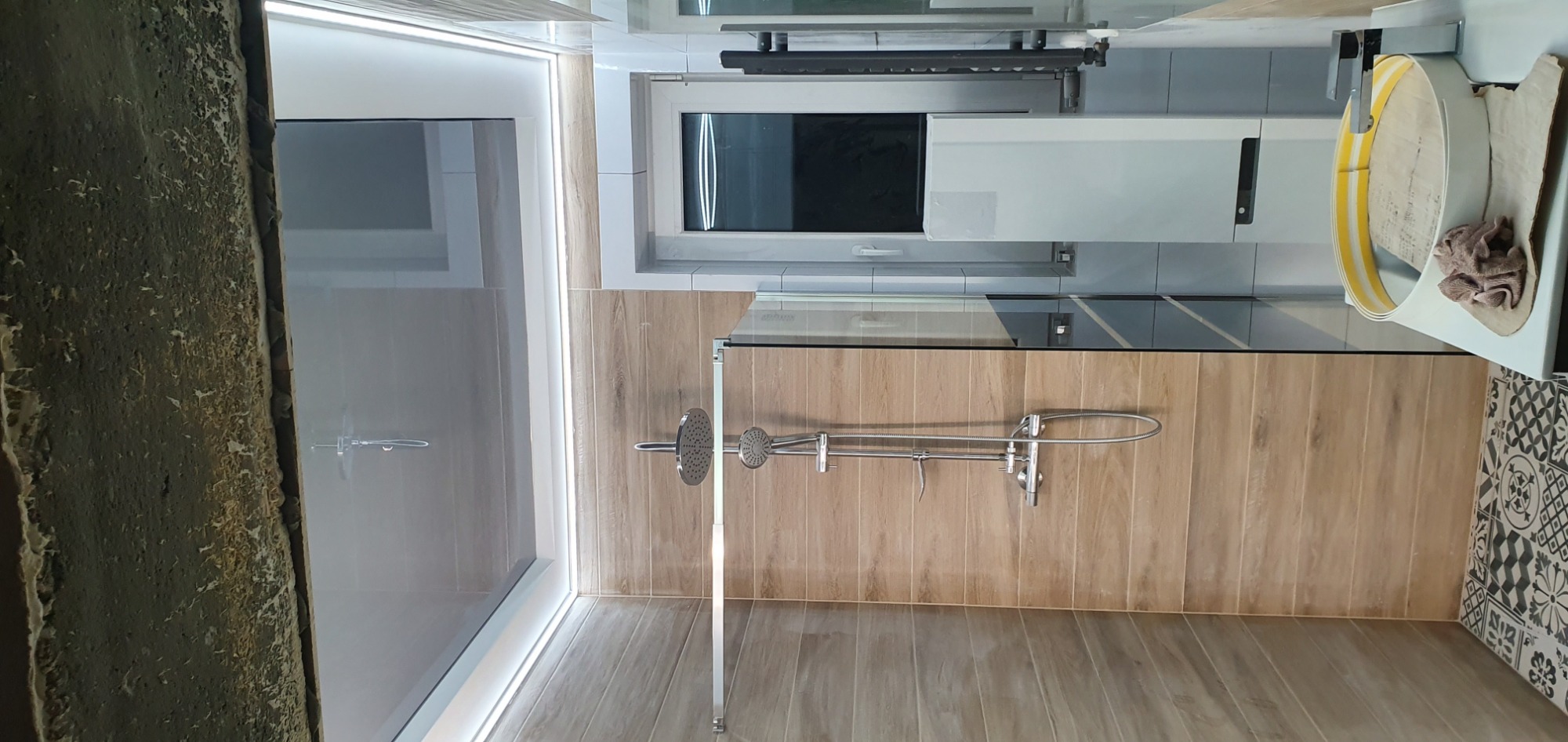 Szklana kabina prysznicowa z deszczownicą i słuchawką, obok umywalka z żółtą taśmą zabezpieczającą, wykończona płytkami imitującymi drewno i czarno-białą mozaiką.