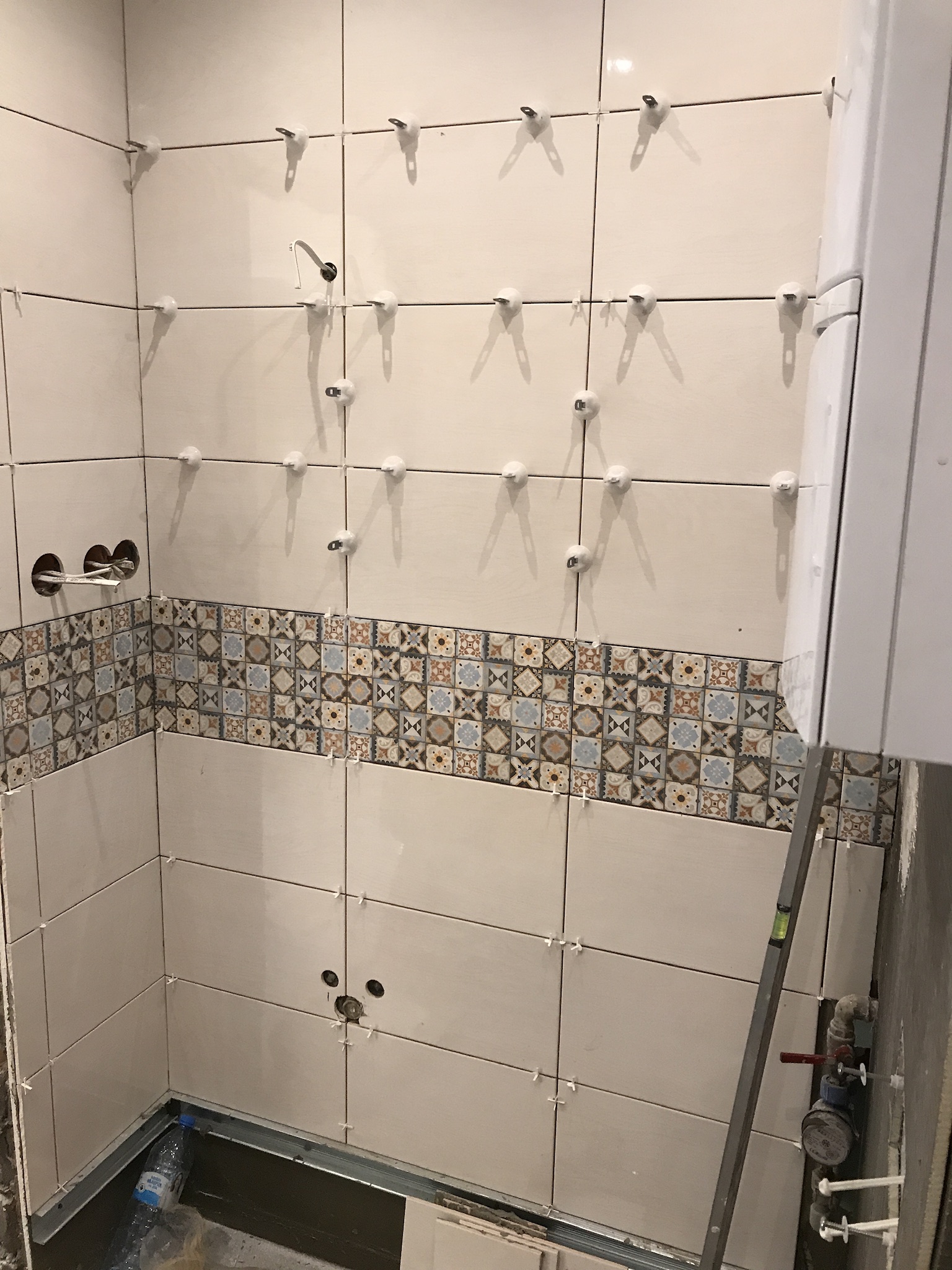 W trakcie układania płytek w łazience; widoczne białe płytki z dekoracyjnym pasem mozaiki, krzyżyki dystansowe, poziomica oparta o ścianę, oraz otwory na armaturę.