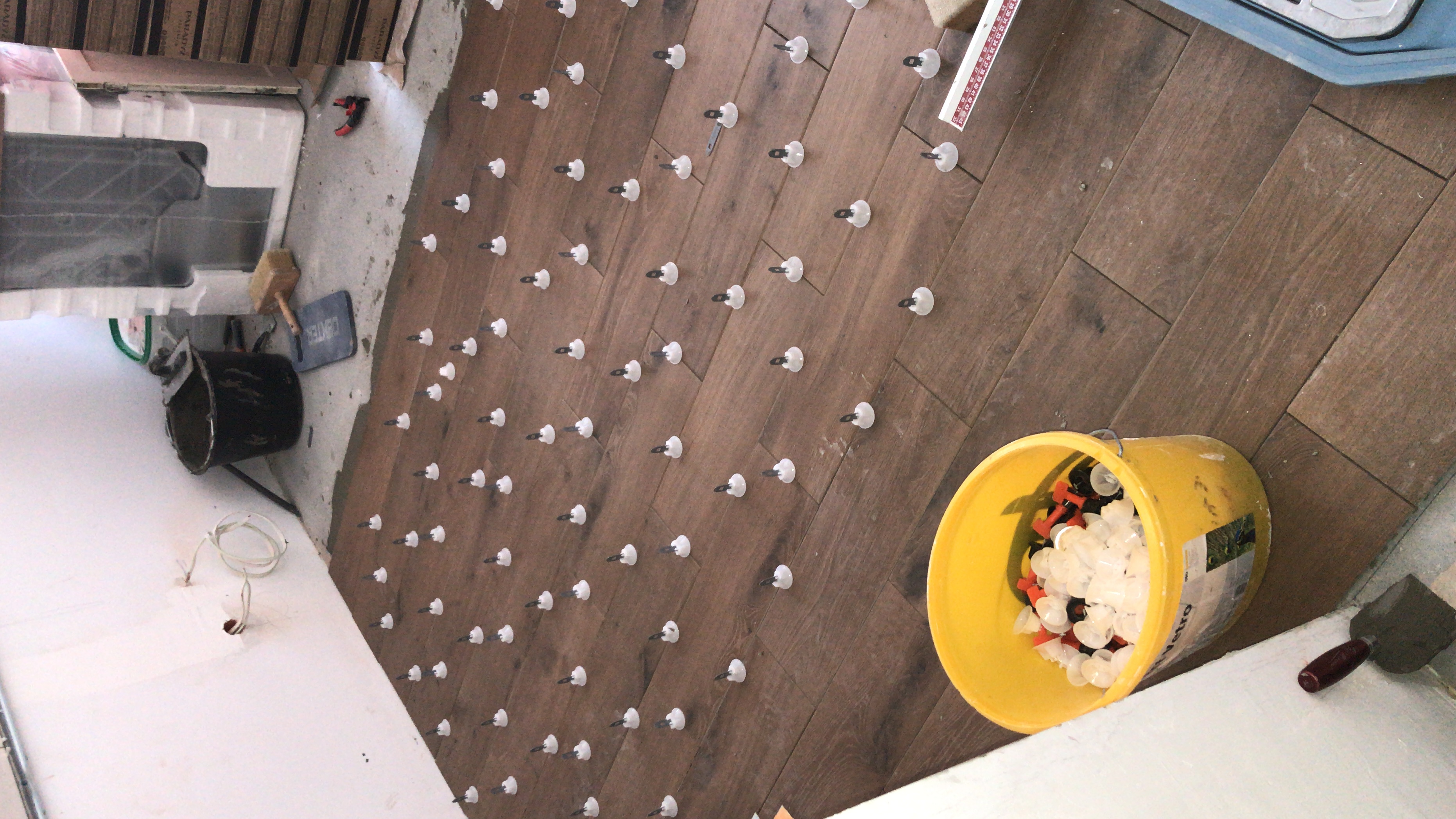 Układanie brązowych płytek imitujących drewno z użyciem klipsów poziomujących; żółte wiadro z klipsami i klinami w rogu pomieszczenia.