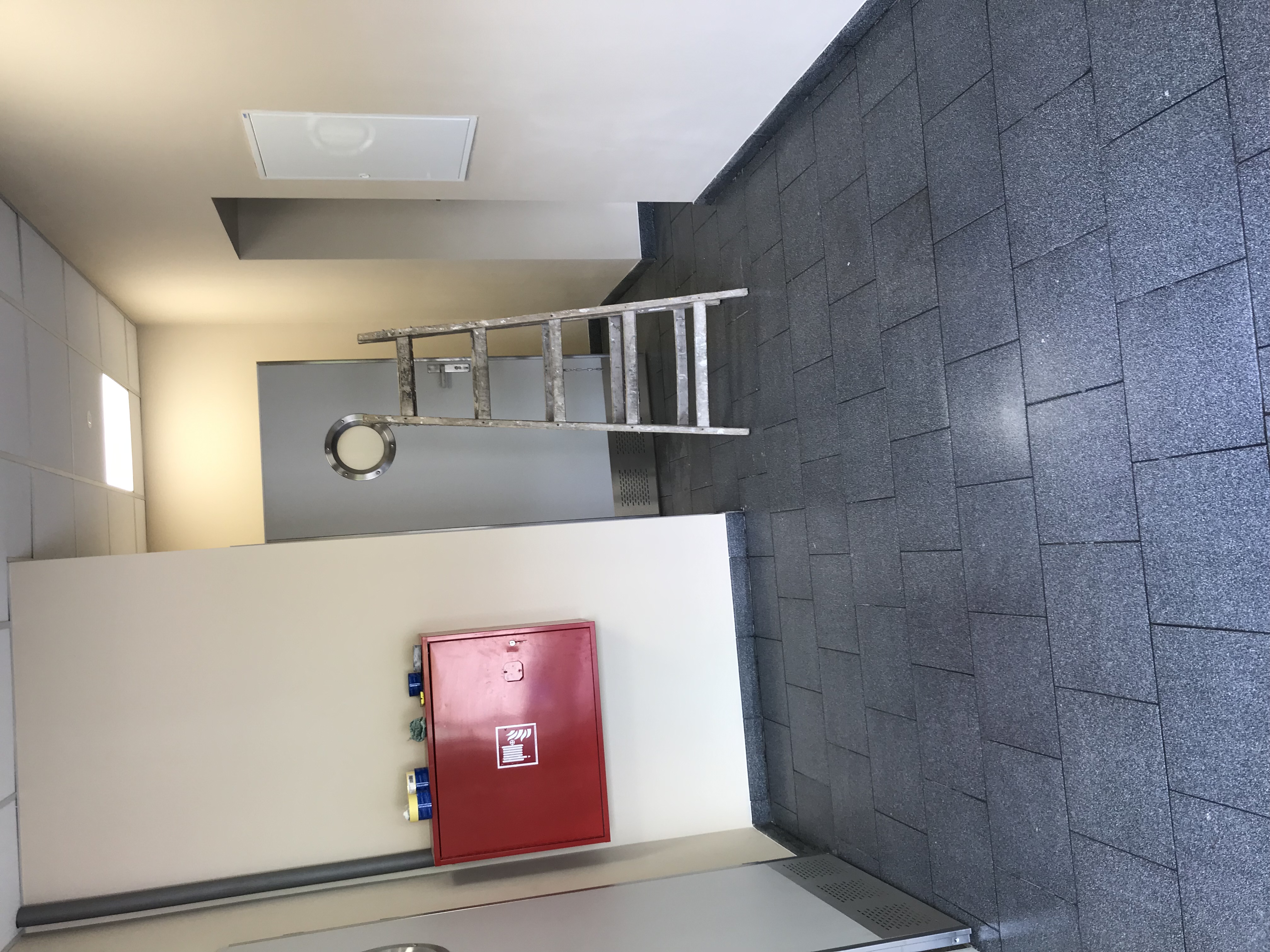 Korytarz wykończony ciemnoszarą, drobną mozaiką ceramiczną na ścianie i podłodze, z aluminiową drabiną opartą o ścianę, czerwoną szafką hydrantową i szarą metalową ścianą z okrągłym okienkiem.