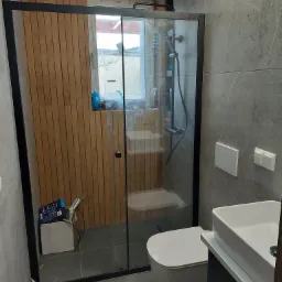 Nowoczesna łazienka z kabiną prysznicową typu walk-in z czarnymi profilami, drewnianą boazerią, szarą płytką imitującą marmur, podwieszaną miską WC i umywalką nablatową.