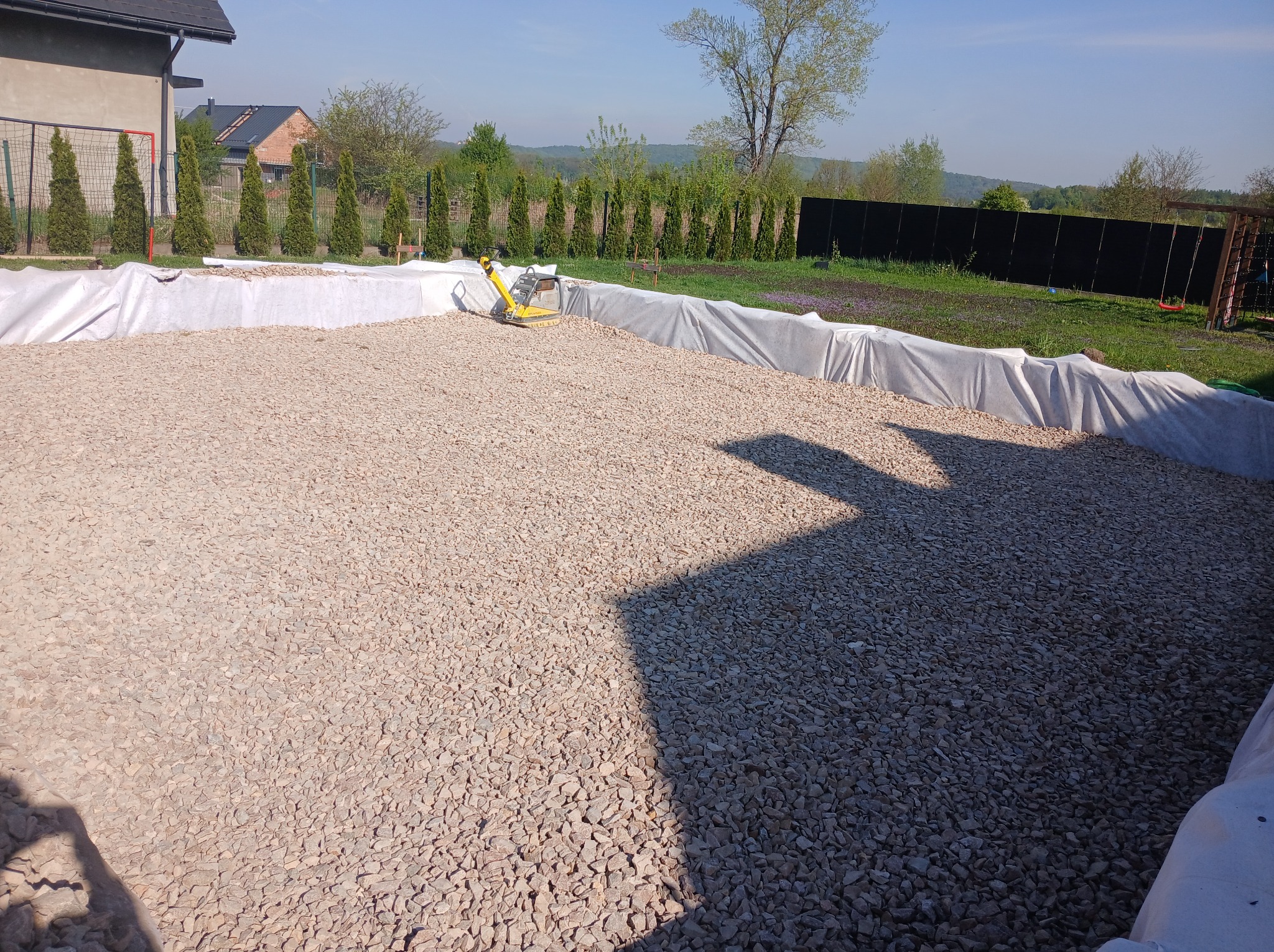Obszar z nasypem z kamienia, przygotowany pod utwardzenie. W tle widoczny zagęszczarka wibracyjna, drzewa iglaste i budynek mieszkalny.