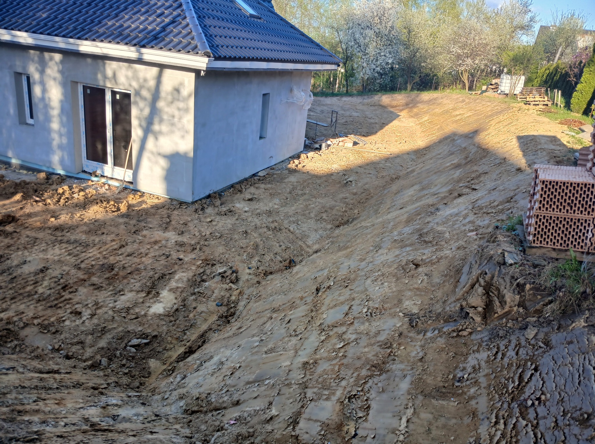 Nierówny teren po niwelacji przy budynku z ciemnym dachem, cegły na skraju. Widoczne ślady opon na ziemi. W tle drzewa z białymi kwiatami.
