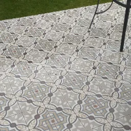 Wzór geometryczny na ułożonej kostce brukowej w odcieniach szarości, beżu i brązu, widok z góry z fragmentem trawnika i nogami metalowych krzeseł.