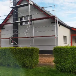 Dwukondygnacyjny dom jednorodzinny w trakcie prac wykończeniowych elewacji, otoczony rusztowaniem, z widocznym kominem i zadbanym trawnikiem z tujami.