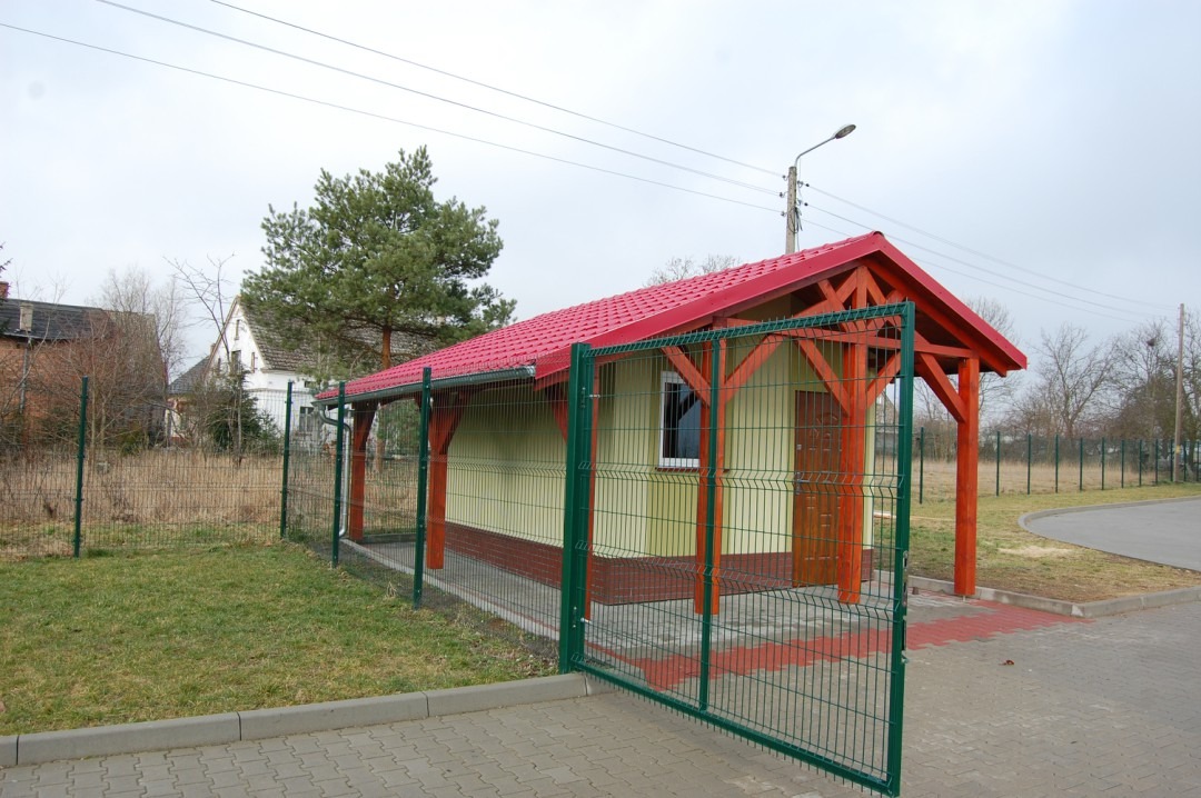 Niewielki budynek gospodarczy z czerwonym dachem i drewnianą konstrukcją, ogrodzony zieloną siatką, z otwartą bramką z kostki brukowej na podjeździe.