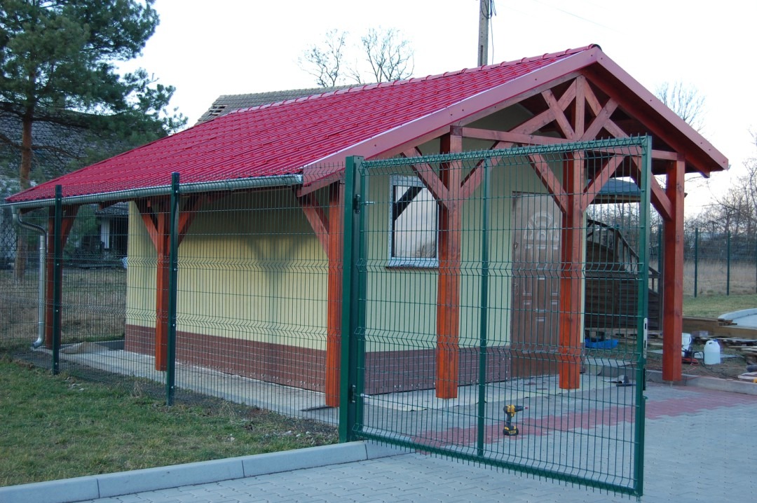 Nowo wybudowany budynek gospodarczy z czerwonym dachem z blachy trapezowej, obity beżową blachą z brązowym cokołem, ogrodzony zieloną siatką panelową z otwartą furtką, na kostce brukowej,...