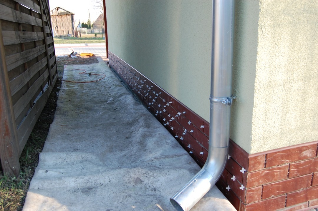 Montaż paneli elewacyjnych imitujących cegłę na cokole budynku, widoczny fragment rynny spustowej i krzyżyki dystansowe między panelami.