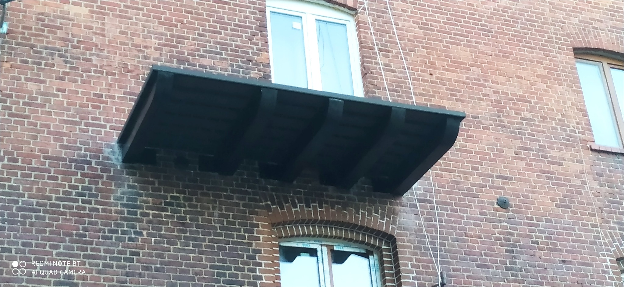 Ciemny, drewniany daszek nad oknem na ceglanej ścianie budynku, widoczne okna z białymi ramami i ślady po instalacji.