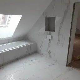 Wnętrze łazienki na poddaszu w trakcie remontu, widoczne białe płytki imitujące marmur na ścianach i podłodze, wanna pod oknem dachowym, przygotowane płyty do montażu z pomarańczowymi oznaczeniami.