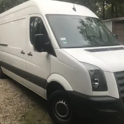 Biały samochód dostawczy typu furgon zaparkowany na żwirowej nawierzchni, widok z przodu lekko z boku, z widoczną naklejką na przedniej szybie.