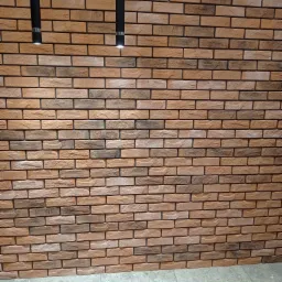 Ściana wyłożona dekoracyjną cegłą w odcieniach brązu i beżu, oświetlona dwoma wiszącymi, czarnymi lampami tubami z metalowymi akcentami.