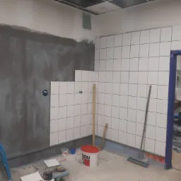 W trakcie prac: Ściana częściowo pokryta szarą zaprawą, a częściowo białymi, kwadratowymi płytkami. Widoczne poziomice, wiadro z zaprawą, narzędzia glazurnicze, oraz niebieska rurka na podłodze.